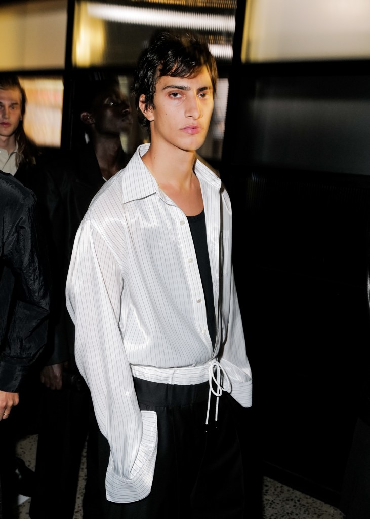 A model with short dark hair wears a tight fitting black shirt under a shiny white, pinstripe button-up shirt.
