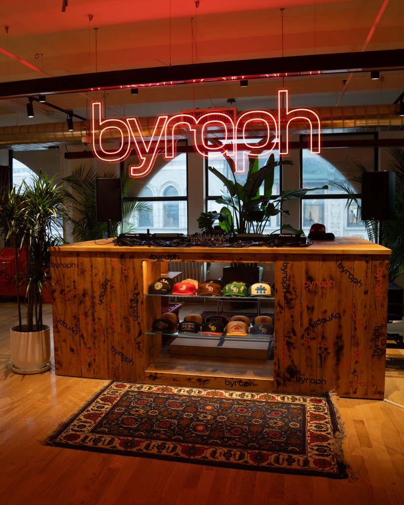 A large neon sign that says "byraph" hangs above a wooden DJ booth displaying collectible ball caps.