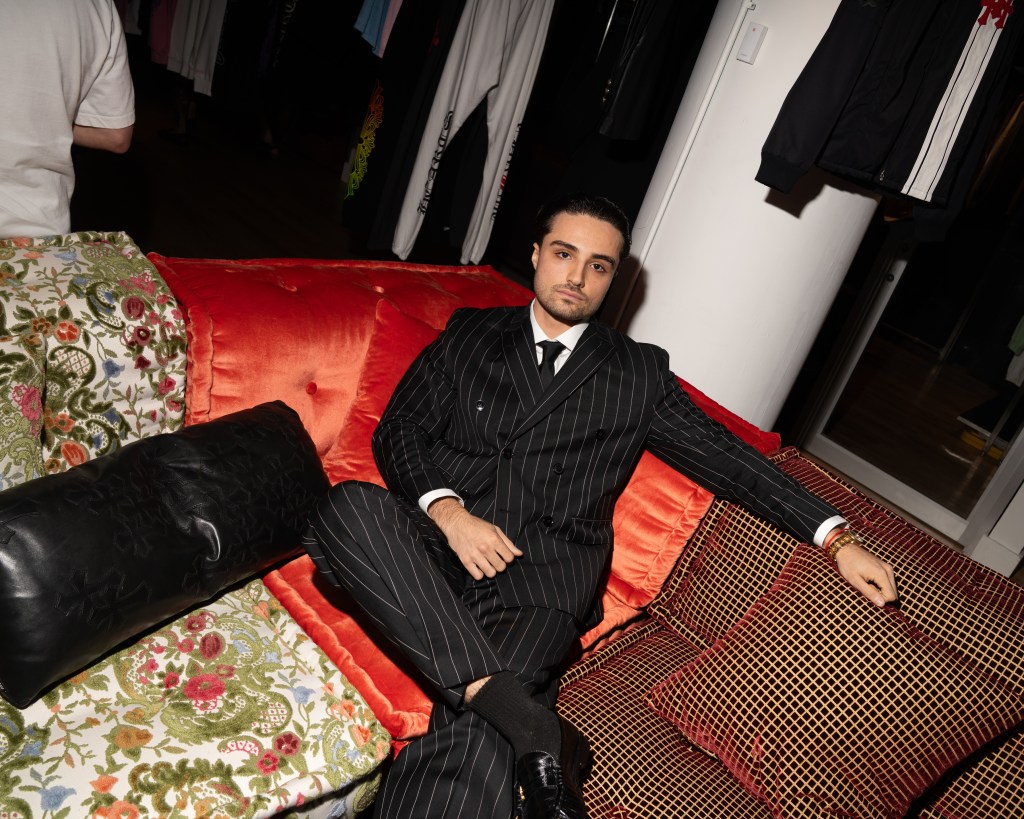 Raphael Badalov sitting in the corner of a red sectional sofa tooling up at the camera with crossed legs.