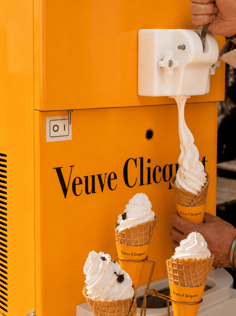 A rendering of a yellow soft serve ice cream machine with a large Veuve Clicquot logo on it, and a hand holding a waffle cone as it fills with ice cream.