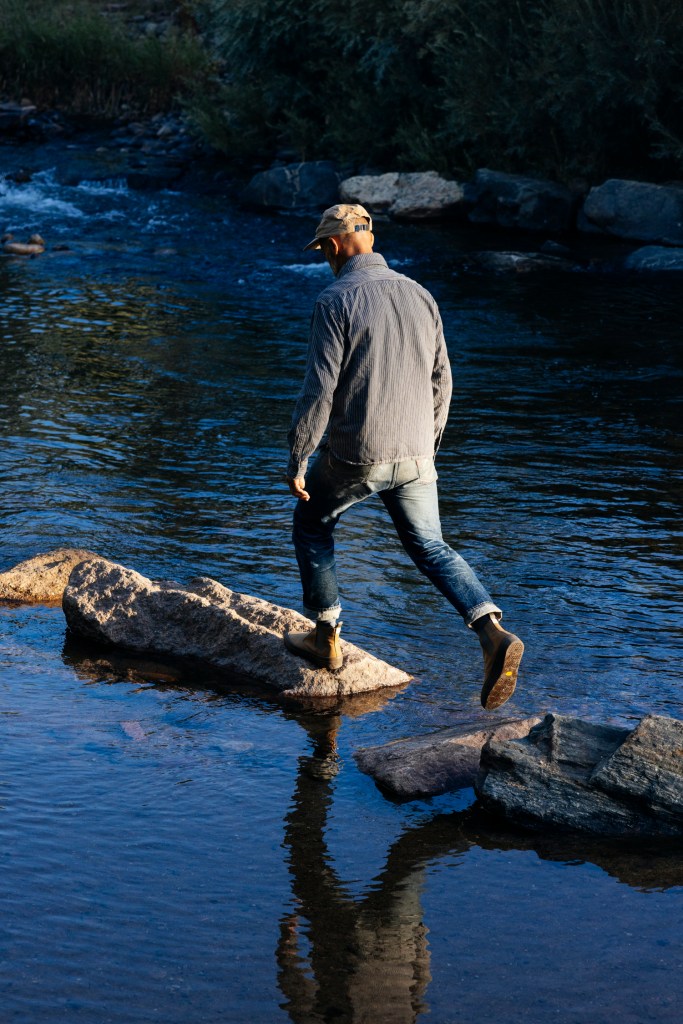 Jeff Johnson wearing a cap, a long sleeve shirt, broken-in jeans and a pair of brown Beyond and Back boots as he walks on large rocks in a body of water.