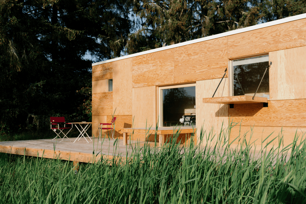 A small wooden, box-like building with square windows and an attached wooden patio with outdoor seating.