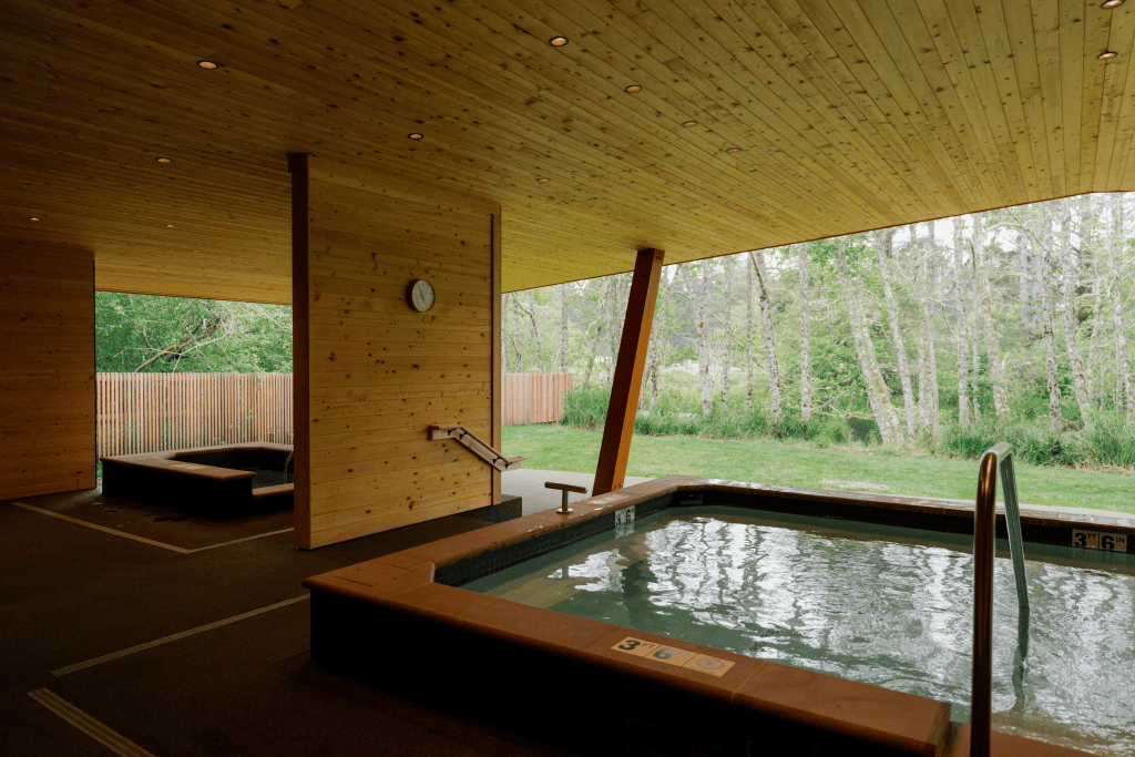 A large outdoor soaking tub looking out onto the woods and grass, with a smaller tub behind a wood wall to the left of frame.