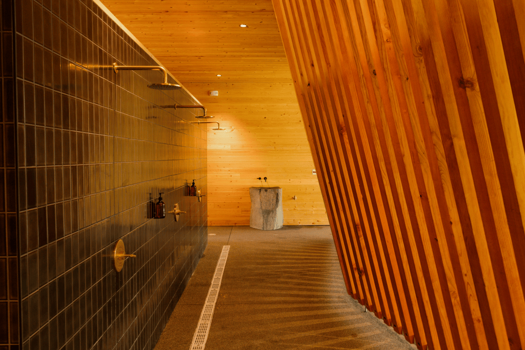 A spa room with angled wooden panels on the right side and a tiled wall of showers on the other, with a sculptural stone sink at the center.
