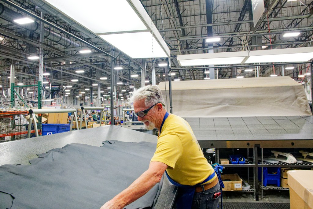 A man wearing a yellow t-shirt leans over a large black piece of leather in a factory.