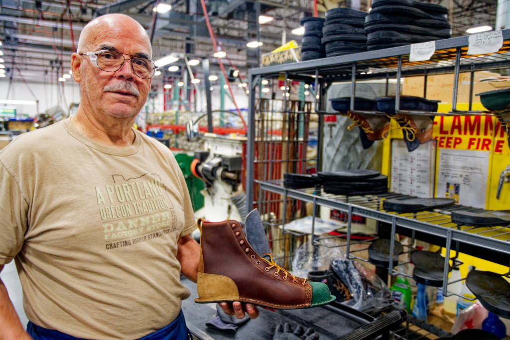 A bald man wearing a tan shirt holds a Danner boot in his hand as he stands in the Danner factory.
