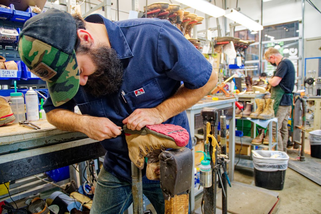 A man with a beard, green hat and navy blue shirt leans over a bench as he removes the outsole of a Danner boot.
