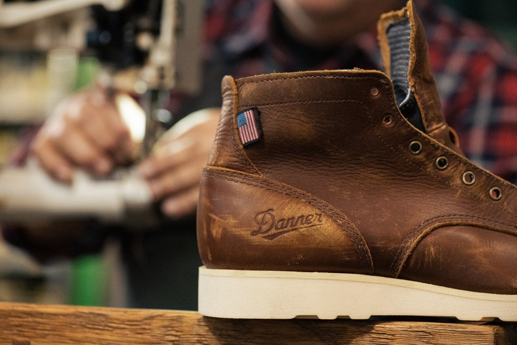 A brown Danner boot sits on a wooden surface with the hands of a shoemaker in the background.