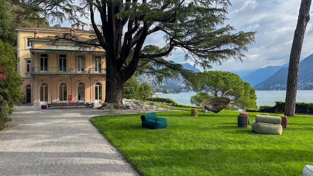 A historic building with modern furniture displayed on the grass with Lake Como to the right of the building.