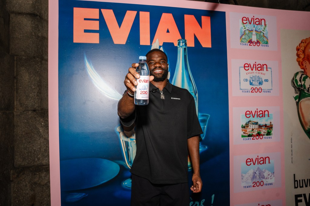 Frances Tiafoe standing in front of archival evian ads, holding a bottle of evian out in front of him.
