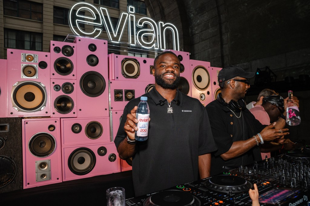 Frances Tiafoe and a DJ standing in front of a booth with light pink speakers and a large evian sign installed behind them.