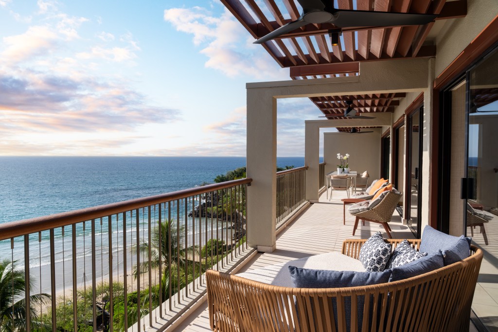 A large lanai at Mauna Kea Resort with sliding glass doors, chairs and tables, looking out onto the ocean below.
