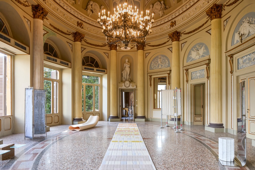 A room at Villa Saporiti with a long rug displayed in the center and other artworks on the sides of the large historic room.
