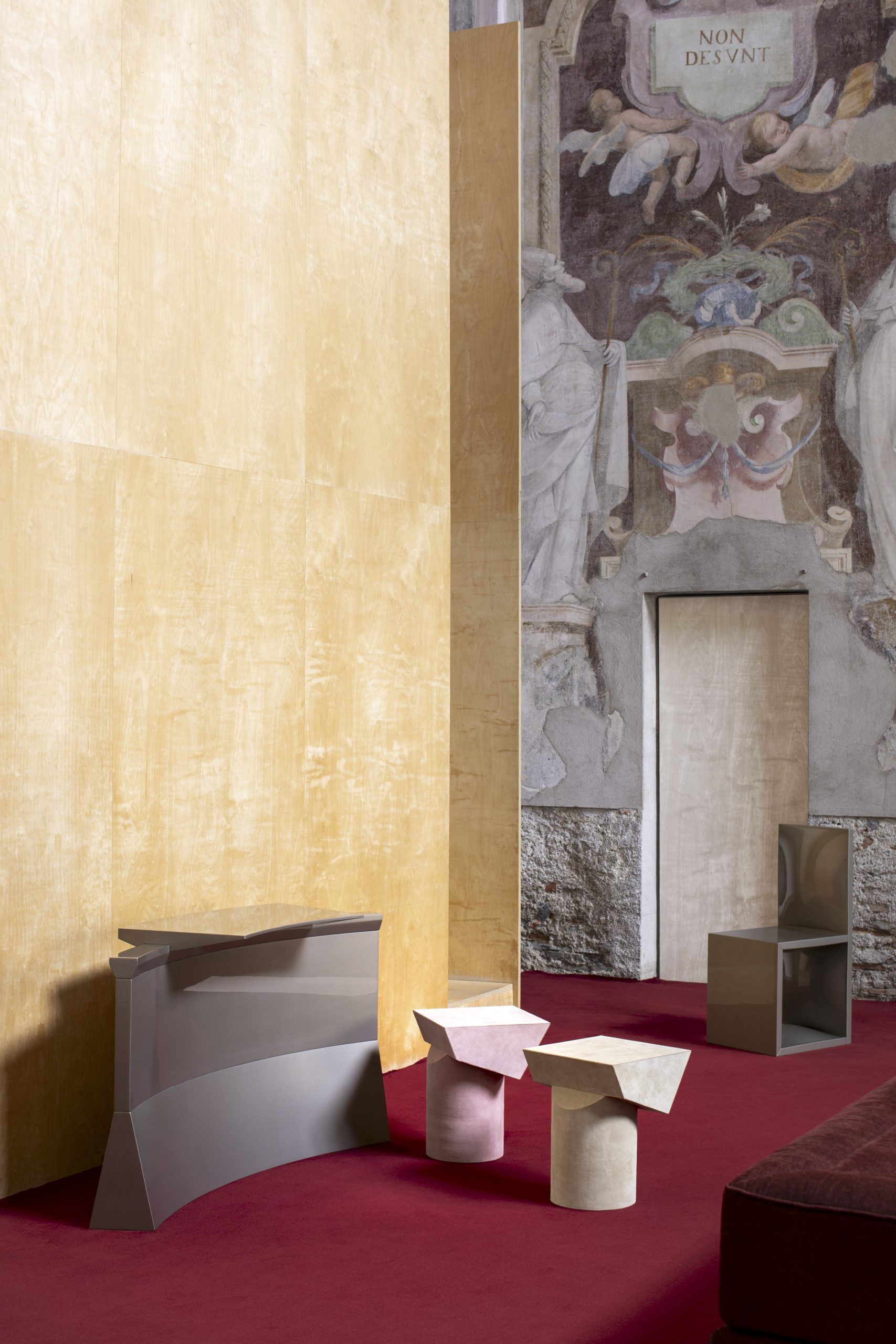 A tall light wood panel on a red carpeted floor with several sculptural pieces of furniture displayed.