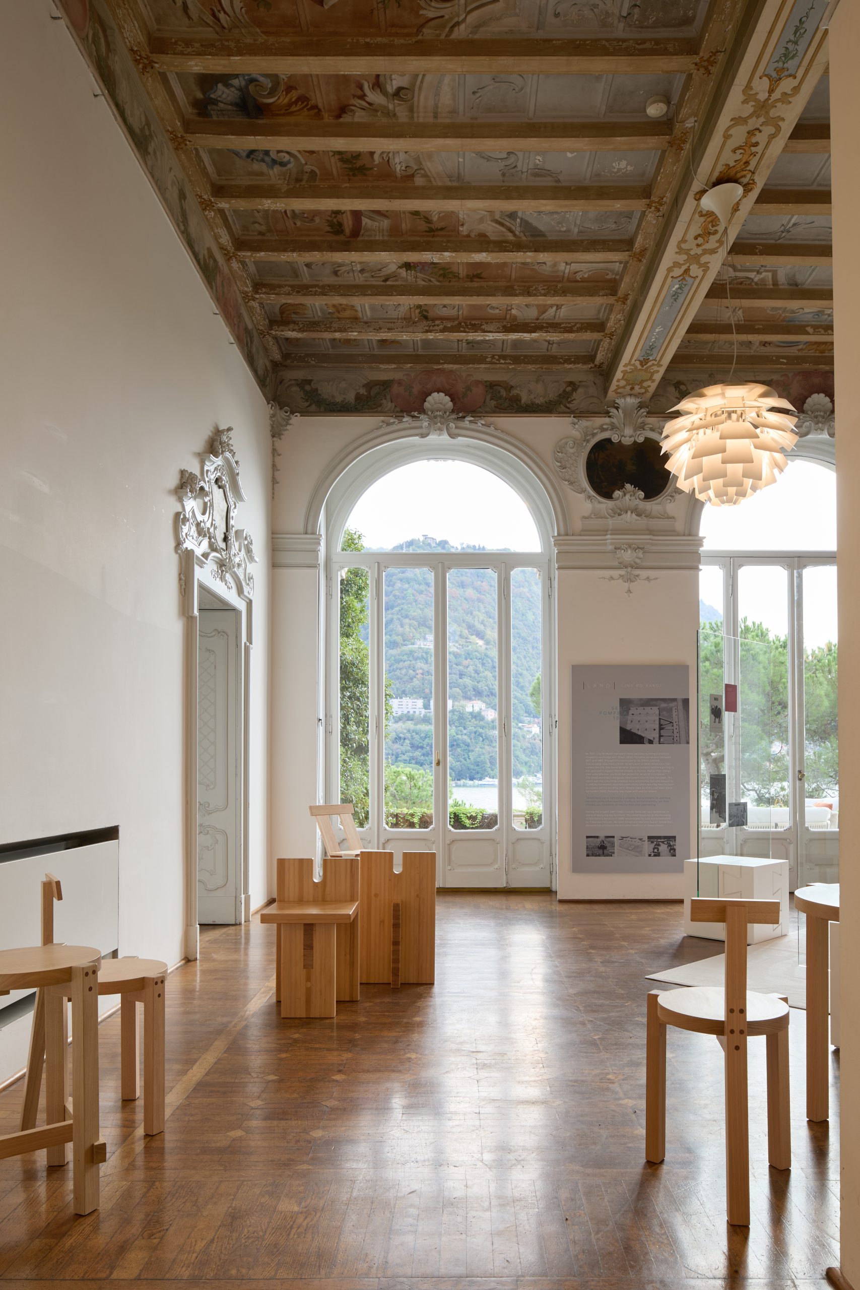 An ornate Italian room filled with simple wooden furniture displayed and arched windows looking out onto Lake Como.