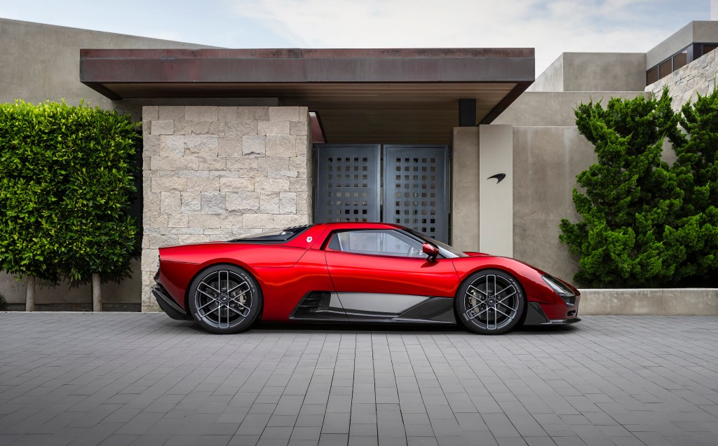 A red McLaren McL 6GT shot from a side profile view, parked in front of a modern tan house.