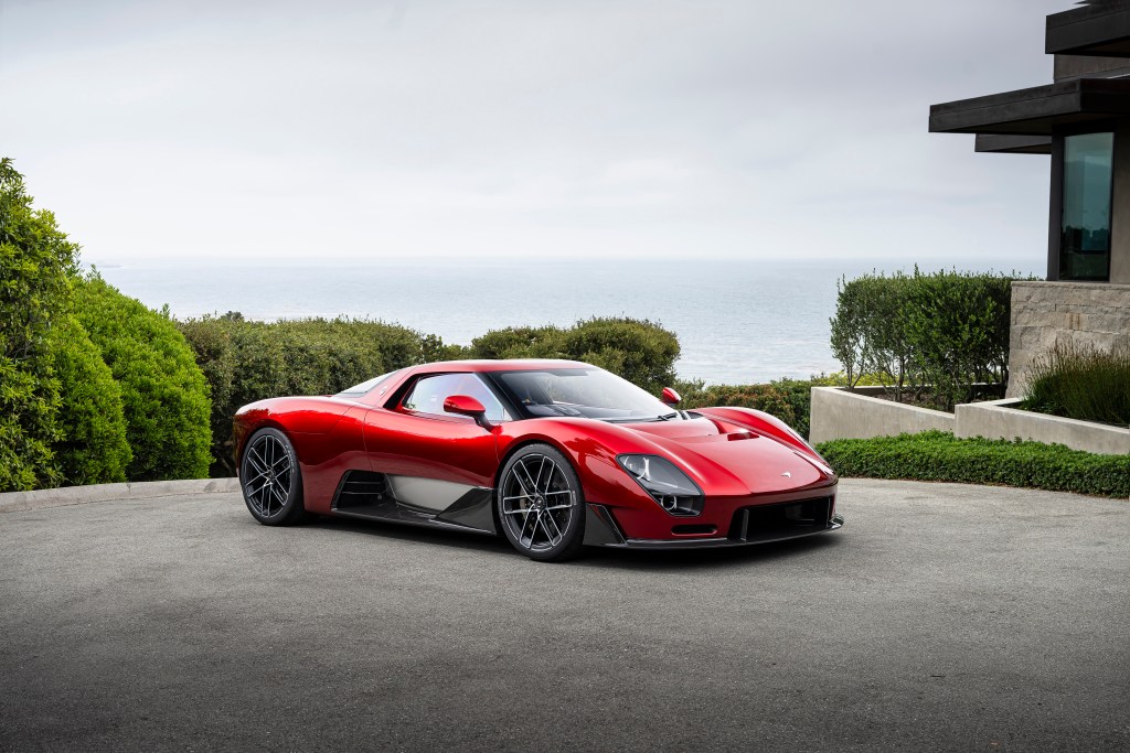 A red McLaren McL 6GT shot at a 3/4 angle, parked in a driveway with the Monterey Bay behind it.