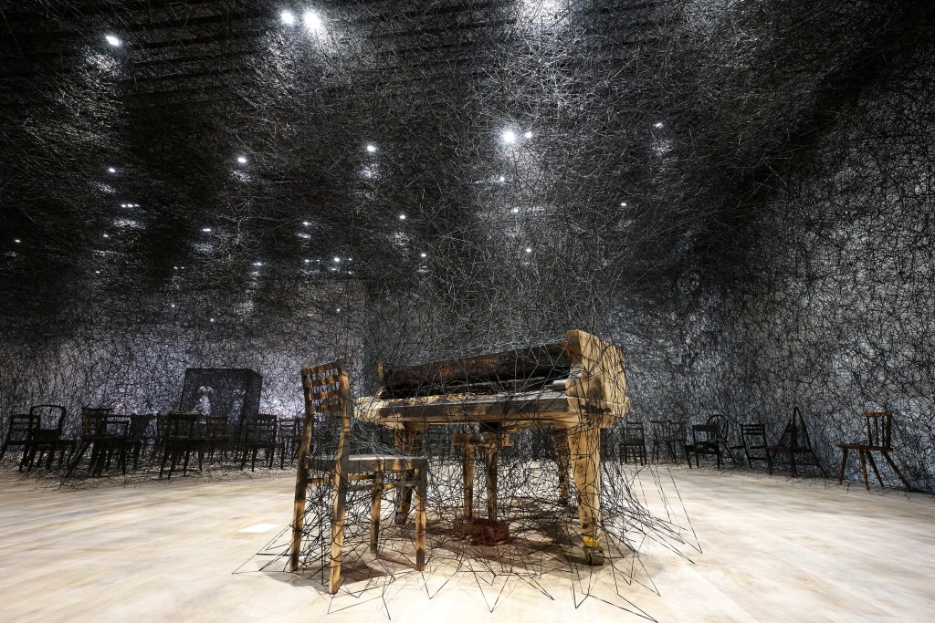A haunting gallery installation of a grand piano and black chairs, all covered in layers of black thread.
