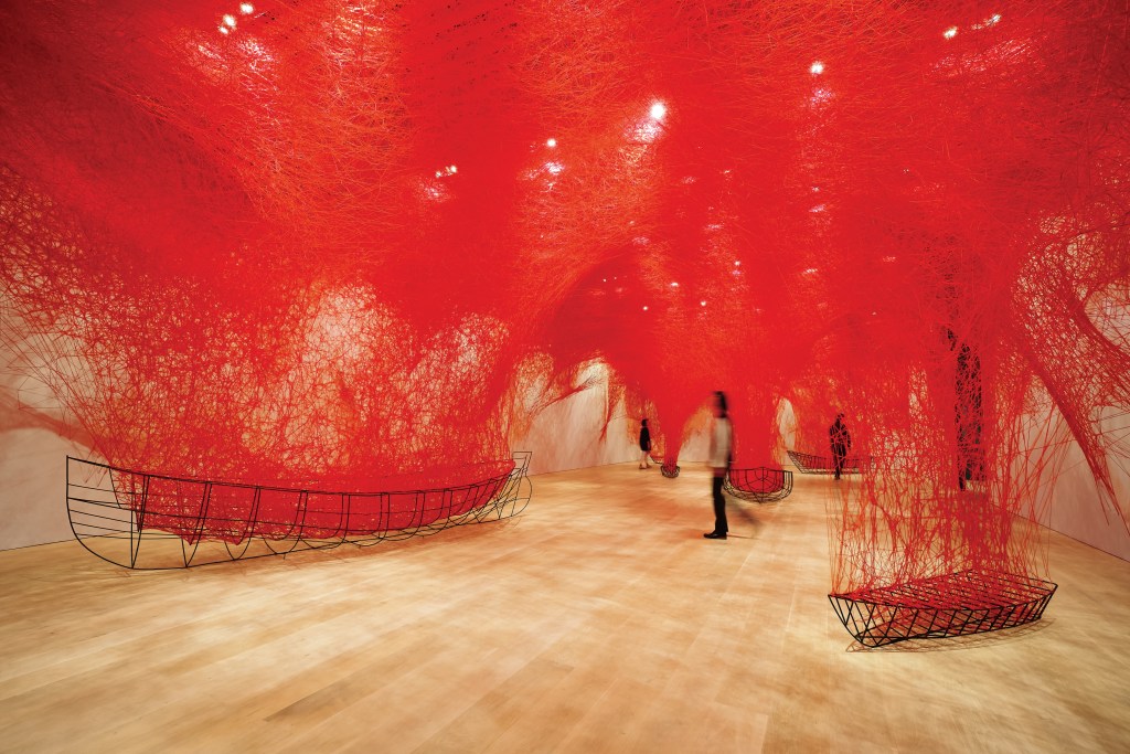 A wood-floored gallery room covered in red threads and people walking through the space.