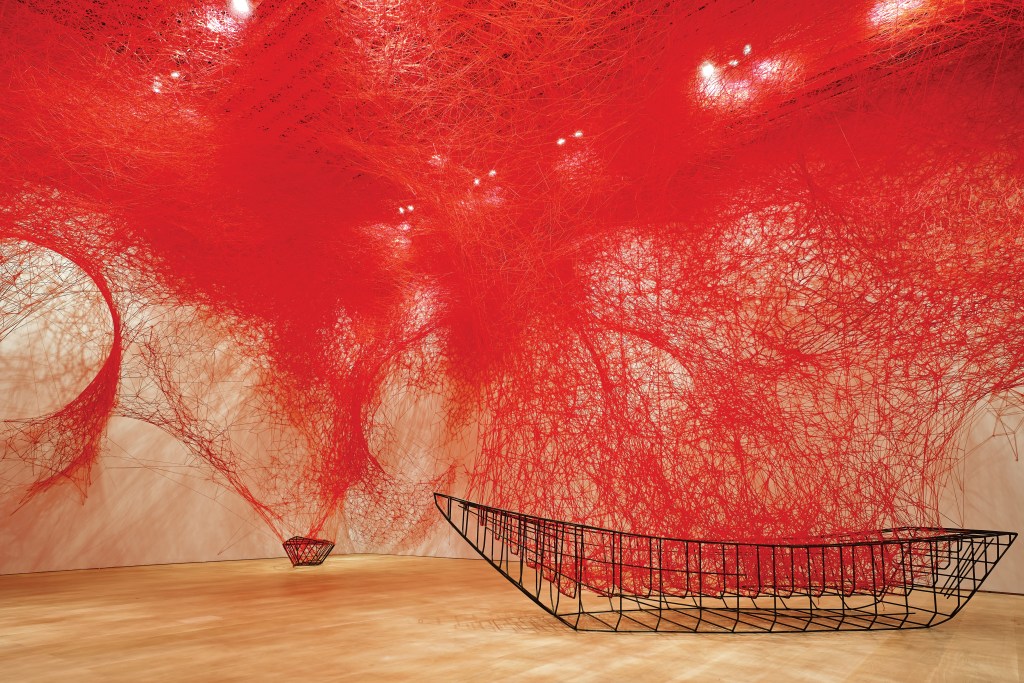 A wood-floored gallery room covered in red threads white a black wire ship shape at the bottom right of the frame.