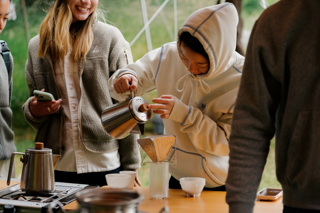 A tight shot of a group of people huddling around an outdoor table as one person makes pour over coffee.