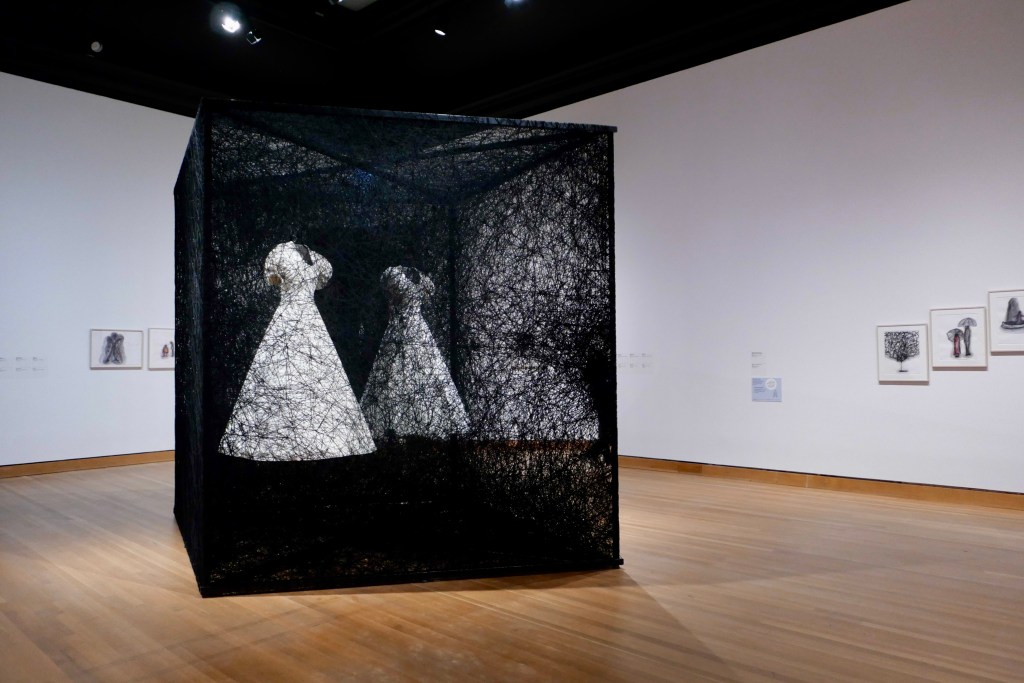 A large installation in a gallery space of a cube made of black thread with a white dress encased inside.