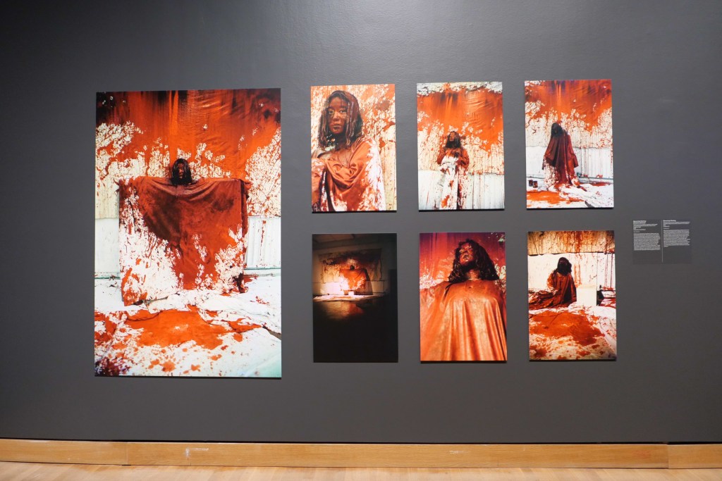 A black gallery wall in a museum with a series of photographs depicting Chiharu Shiota covered in red, blood-like material.