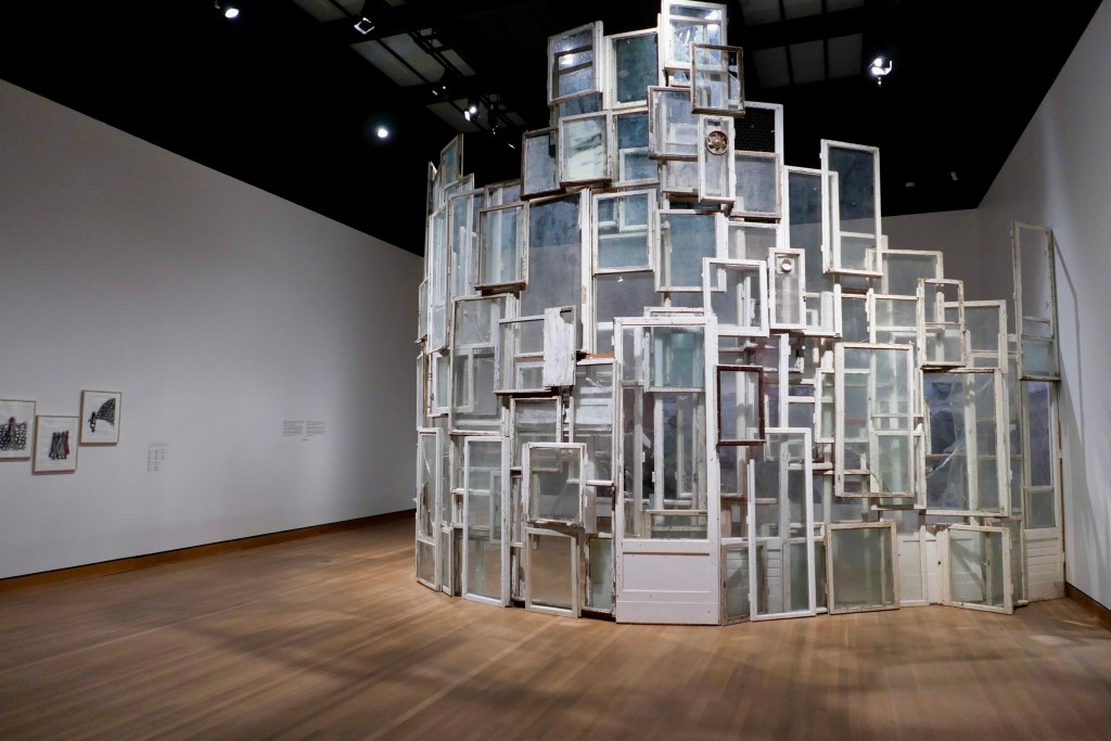 A stack of white framed windows of various sizes stacked up to the ceiling in a large gallery room.