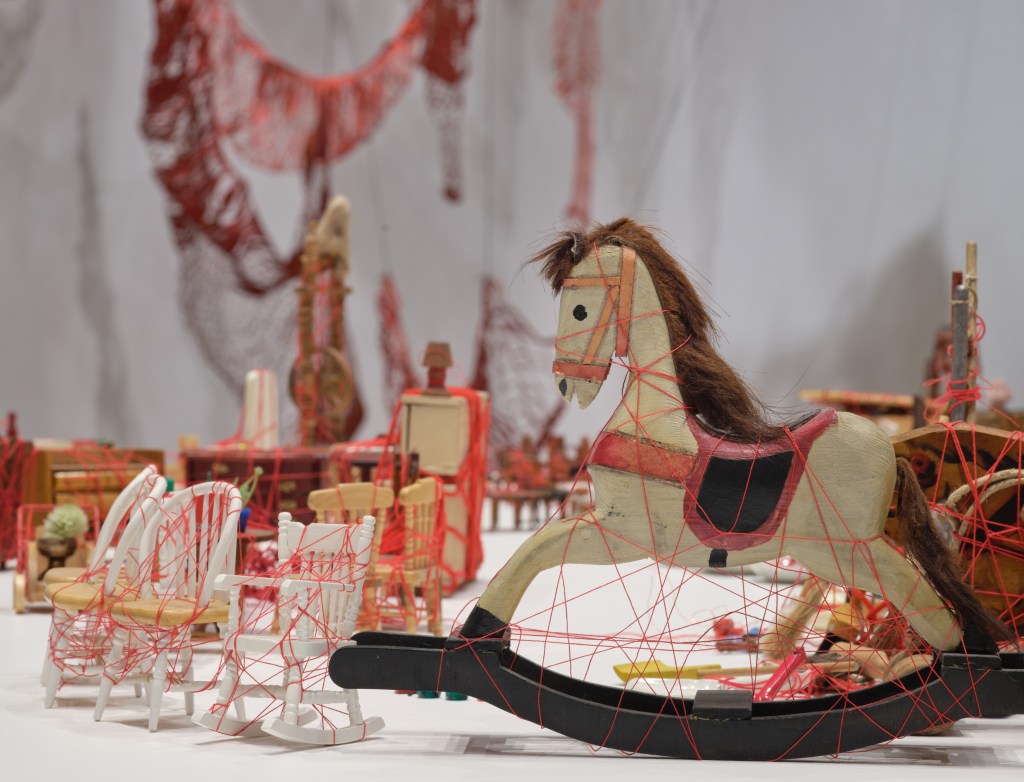 A detail of an installation of wooden childhood objects like a miniature rocking horse and dollhouse items wrapped in red threads.