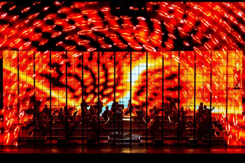 An orchestra plays behind a transparent LED screen with a vibrant red and orange semi-circular design being projected onto it.