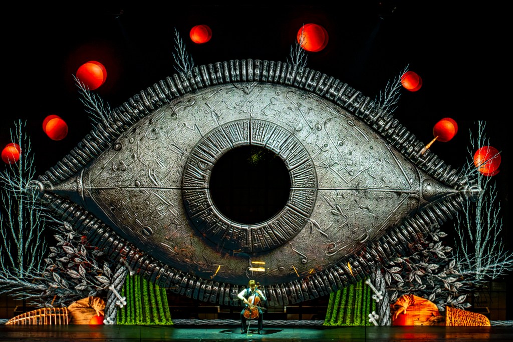 A man playing cello at the center of the stage with a large screen behind him depicting an image of an oversized, stylized eye with other characters surrounding it.