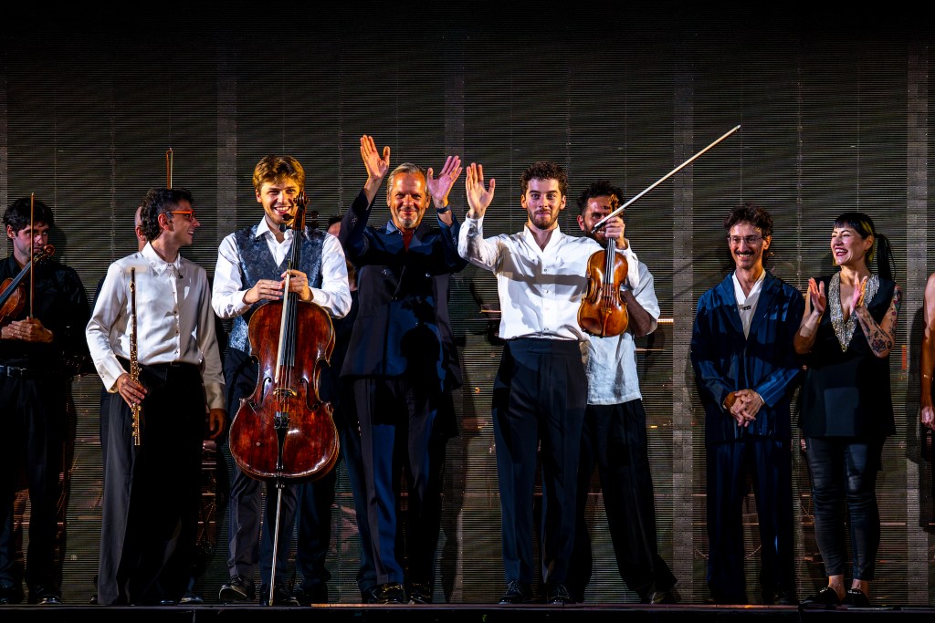 A cellist, a man in a suit, and a violinist stand on stage flanked by other musicians. 