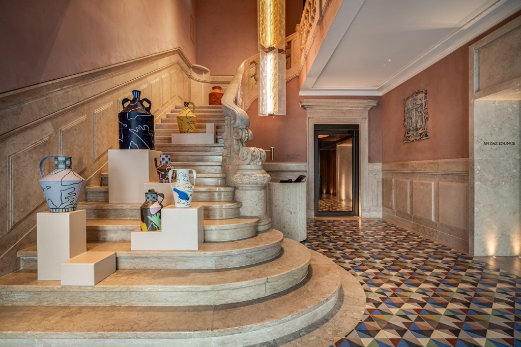 A part of the Andaz Lisbon lobby with a reception desk on the right, and a historic marble staircase with various ceramic vessels displayed on the lower steps.