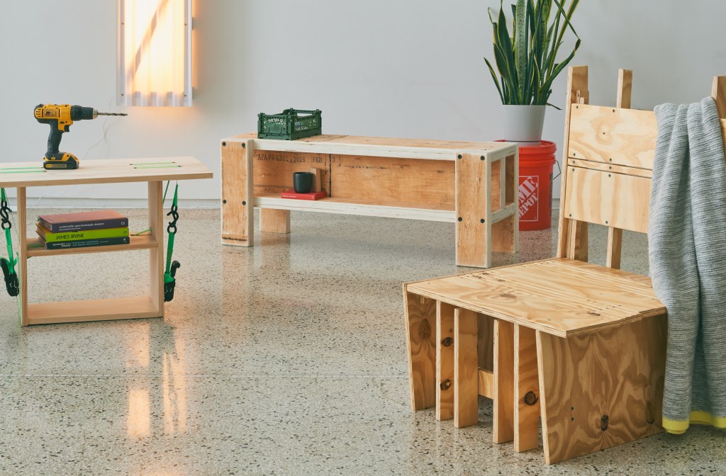 A room with several plywood pieces of furniture, a white wall with a DIY light sconce and an orange bucket with a plant in it.