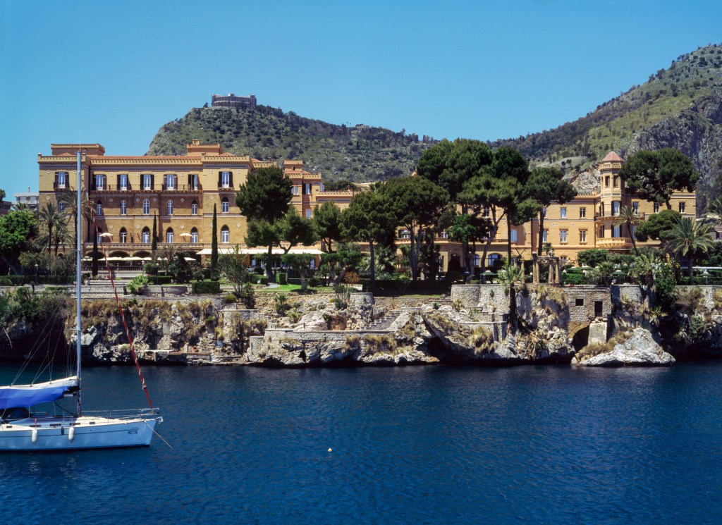 An exterior shot of Villa Igiea, a Sicilian hotel perched on a rocky ledge overlooking a dark blue body of water.