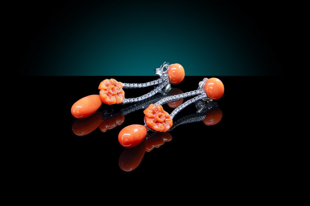 A pair of diamond and ornately carved coral earrings shot on a reflective black surface.