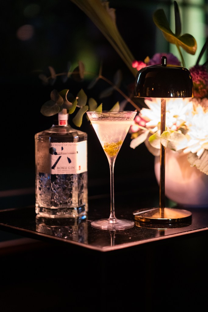 A cocktail and a bottle of Roku Gin in a dark bar with a warm light illuminating the scene.