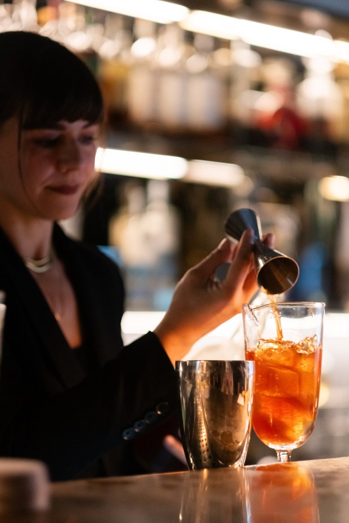 Julia Momose pouring a cocktail at a bar with soft light in the background.