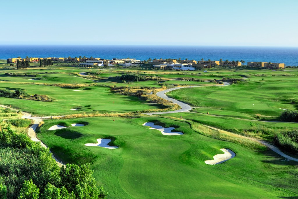 A sprawling green golf course and a large, low-slung building in front of a blue shoreline.