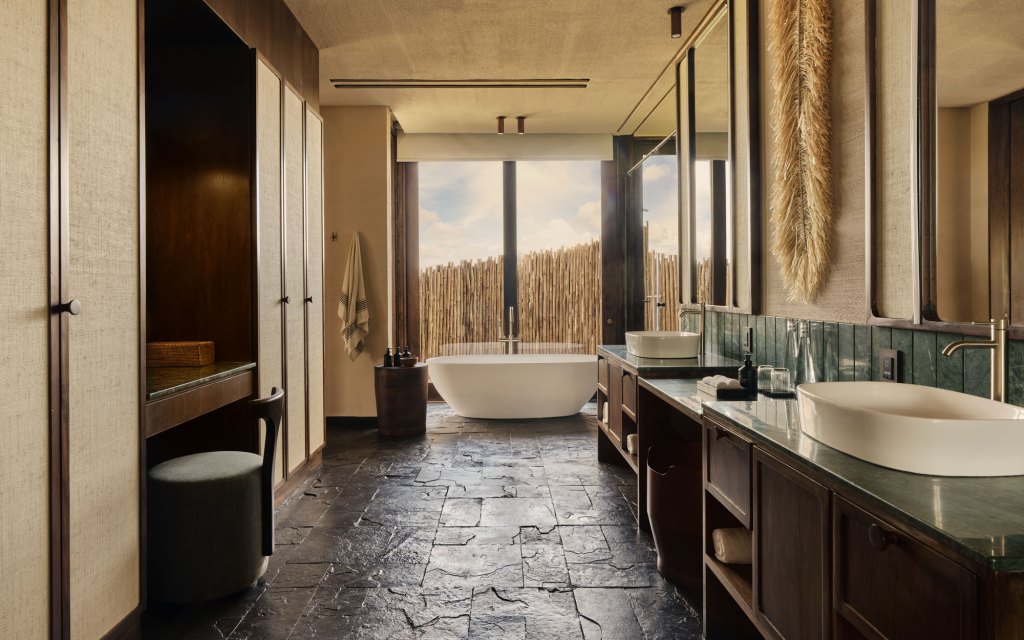 A view looking into the bathroom of a Kitirua Plains Lodge guest suite with closets on the left, double sinks on the right and a large soaking bathtub at the end of the room with full windows looking out.