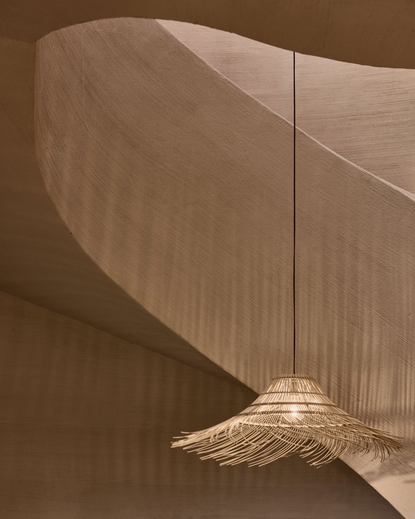 A detail of a woven grass light pendant hanging in front of a curved plaster stairwell. 