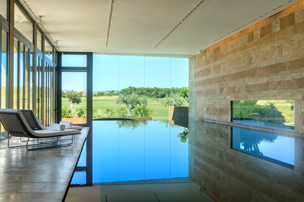 A view from a partially indoor pool, looking out onto green grass, trees and blue sky beyond.