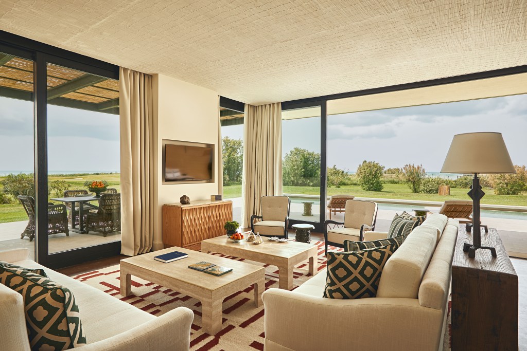 A large living room in a guest suite at Verdura Resort with warm lighting, cream walls and furniture, light wood details and floor-to-ceiling windows that look out onto the sea and golf course. 