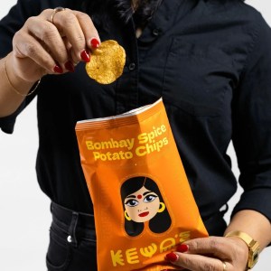 A woman wearing black holds a A bag of Keya's Bombay Spice Potato Chips
