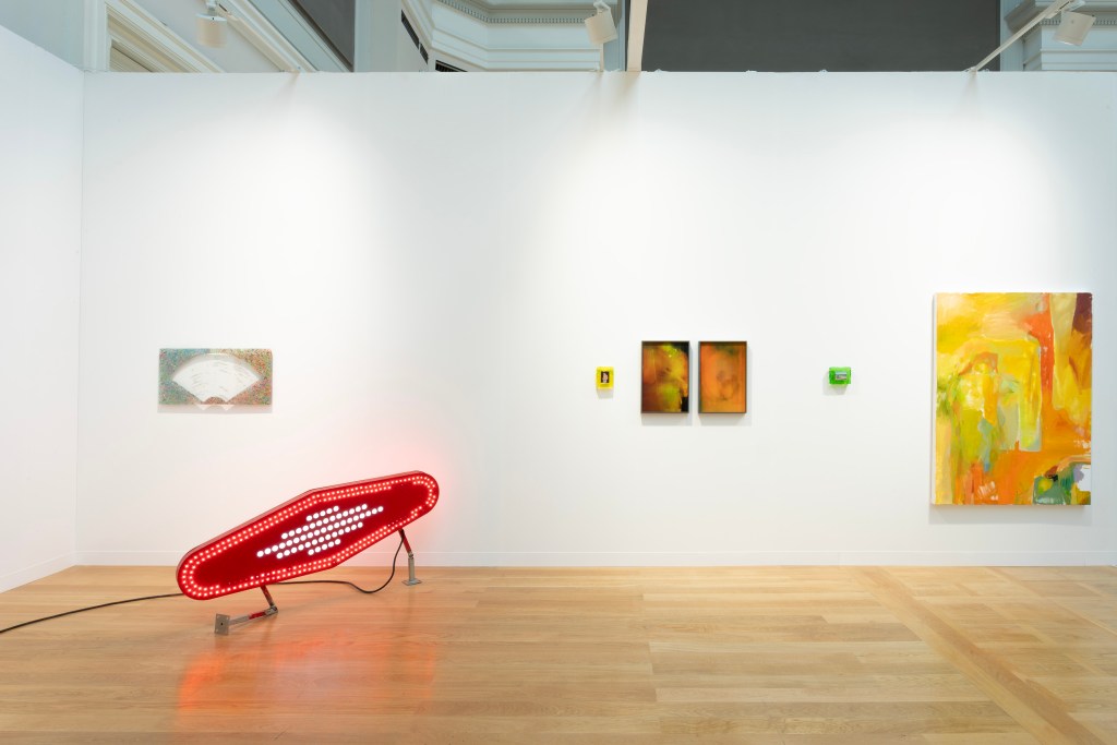 A white gallery room with wood floor showing an illuminated red sculpture on the ground with several hanging artworks on the wall behind it.