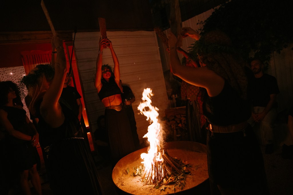 A group of female performers with their arms raised as they stand around a firepit.