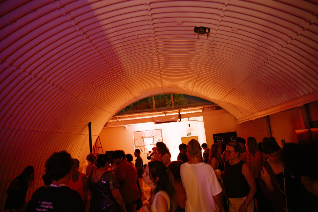 A large group of people standing in a long, dimly light room with a corrugated metal arched ceiling.