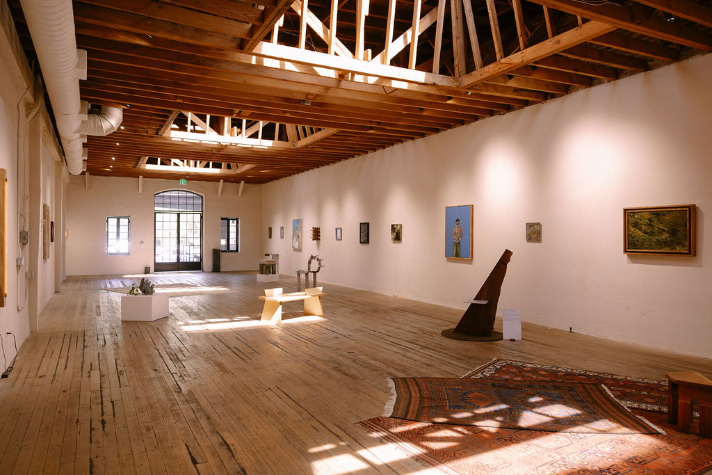 A gallery room with high vaulted wood ceilings and windows at the end of the space with a collection of artworks displayed in the space, with lights from above shining down.