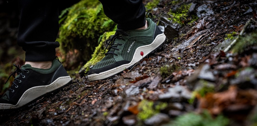 A pair of feet wearing a green pair of Techyak on a mulched surface, shot on an angle.