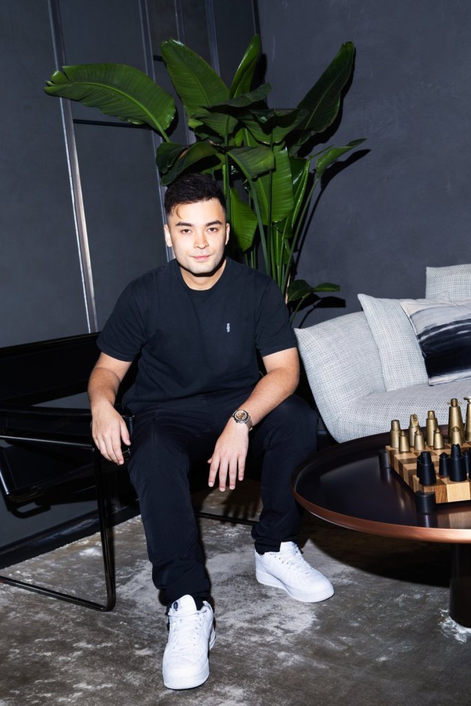 A man wearing a black t-shirt, black pants and white sneakers sits on a bench in a corner with a tall plant behind him.